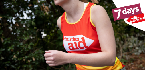 A woman running. There are trees behind her, and she's wearing and red and yellow Christian Aid running vest.