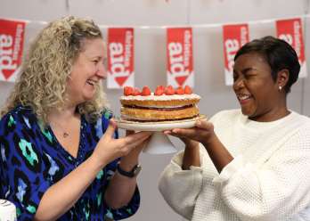 Two women holding a cake. They are both smiling, and there is Christian Aid bunting in the background.