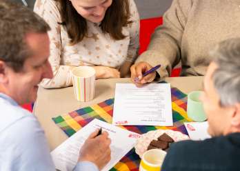 A table displaying brownies and cups of tea. A group of people are leant on the table, filling in a Christian Aid quiz.