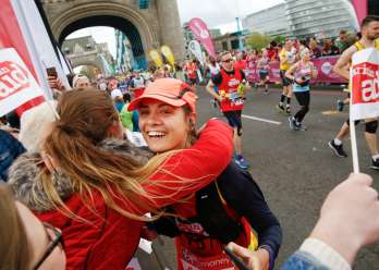 Two women hugging. One is wearing a cap and is running along a marathon track, the other is supporting her from the crowd. There are Christian Aid flags around, and many people are wearing red Christian Aid shirts.